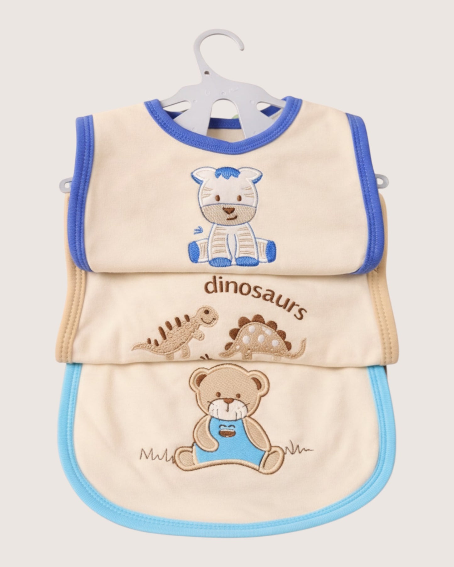 Little Roots pack of 3 cream cotton baby bibs with blue trim, featuring embroidered zebra, dinosaurs, and teddy bear designs.