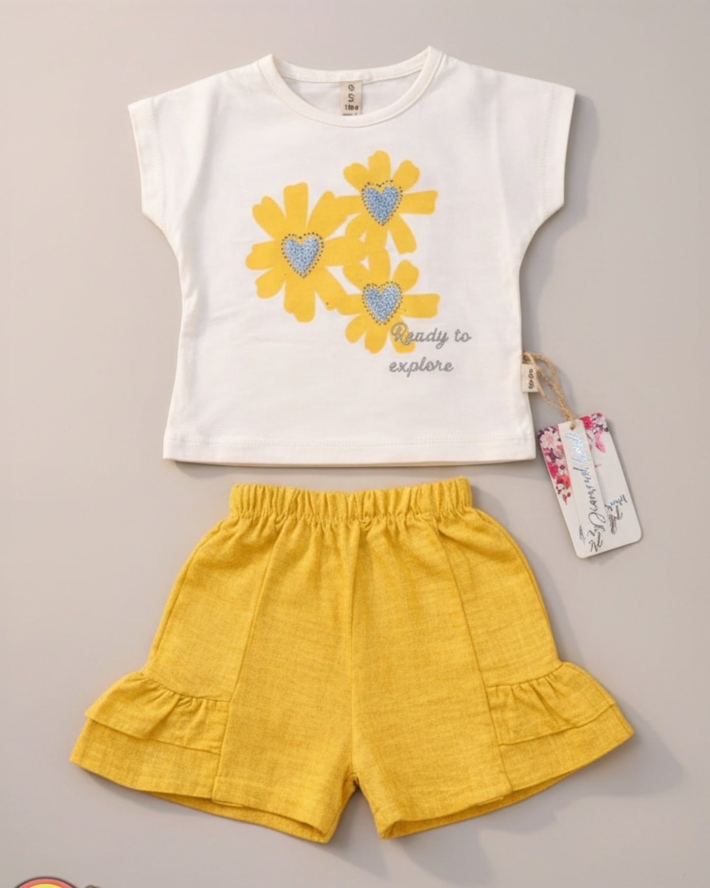 Little Roots Pakistan cream t-shirt with yellow floral print and glitter hearts, paired with yellow ruffle shorts. "Ready to explore" text.